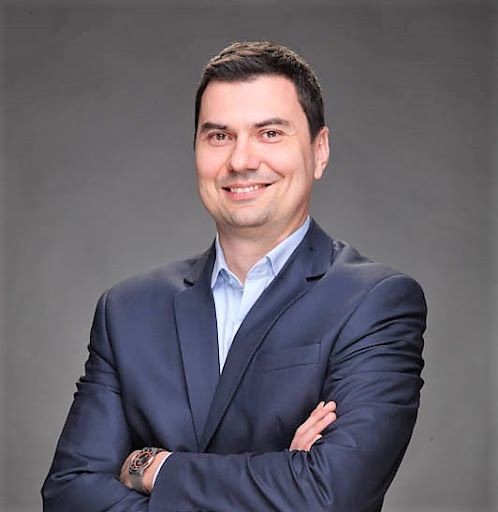 Man in a navy suit with arms crossed, smiling against a gray background.